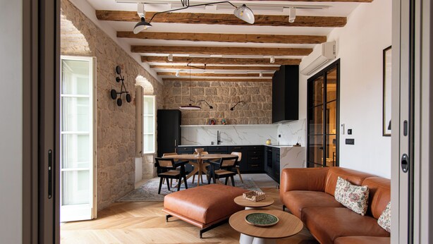A modern living room with a brown leather sofa, a coffee table, and a dining area with a stone wall and wooden beams. - Santa Croce Suite by DuHomes (Dubrovnik)