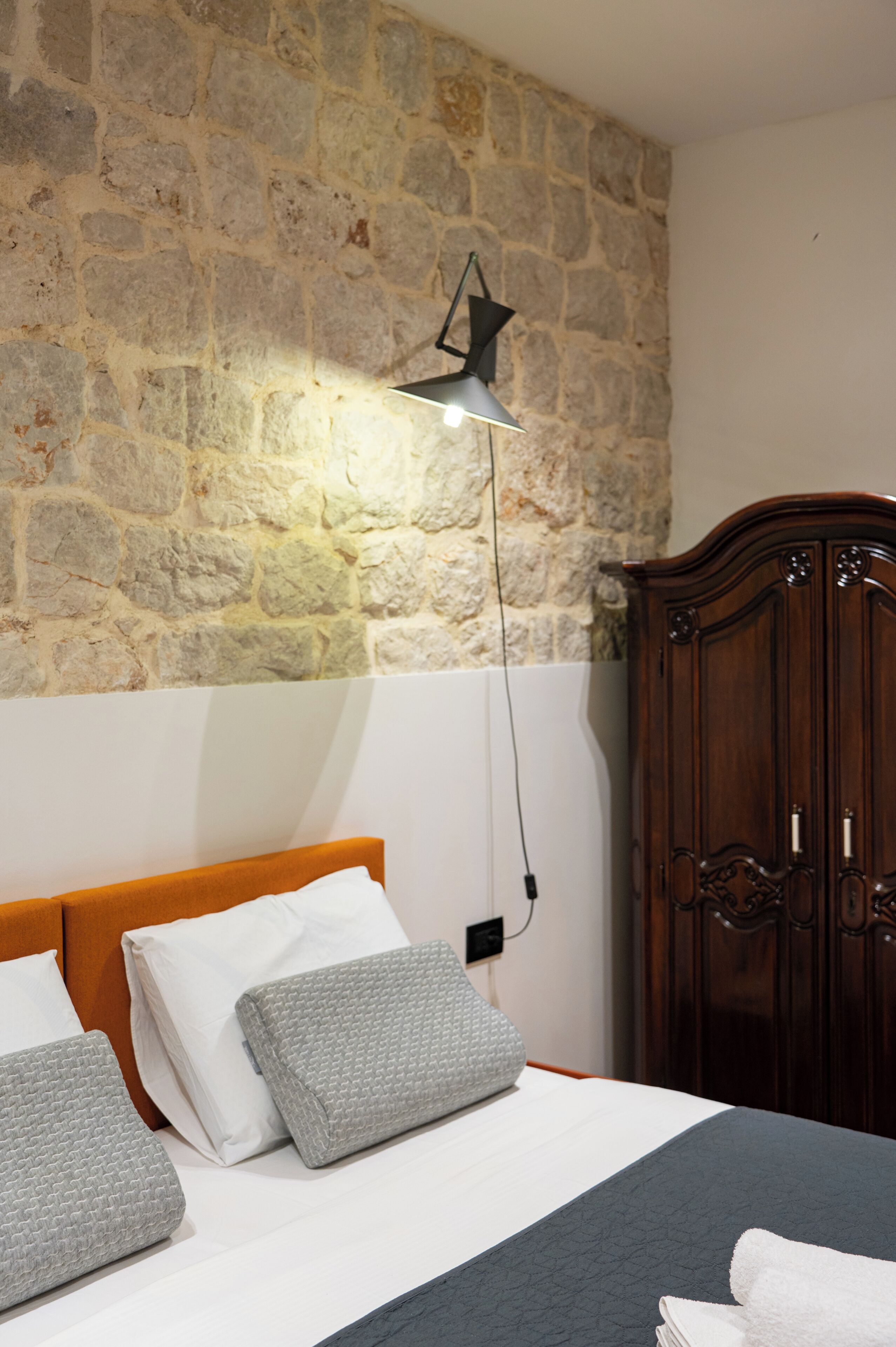 Une chambre avec un mur en pierre, une tête de lit en bois, un lit avec des oreillers, une lampe de chevet et une armoire.