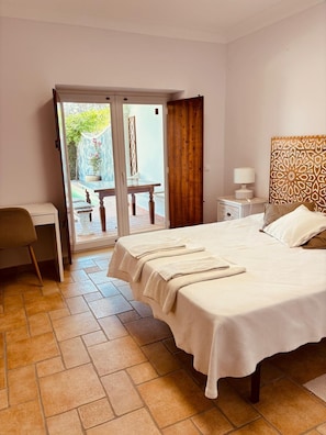 A bedroom with a bed, a desk, a chair, a nightstand, a lamp, and a view of an outdoor patio.