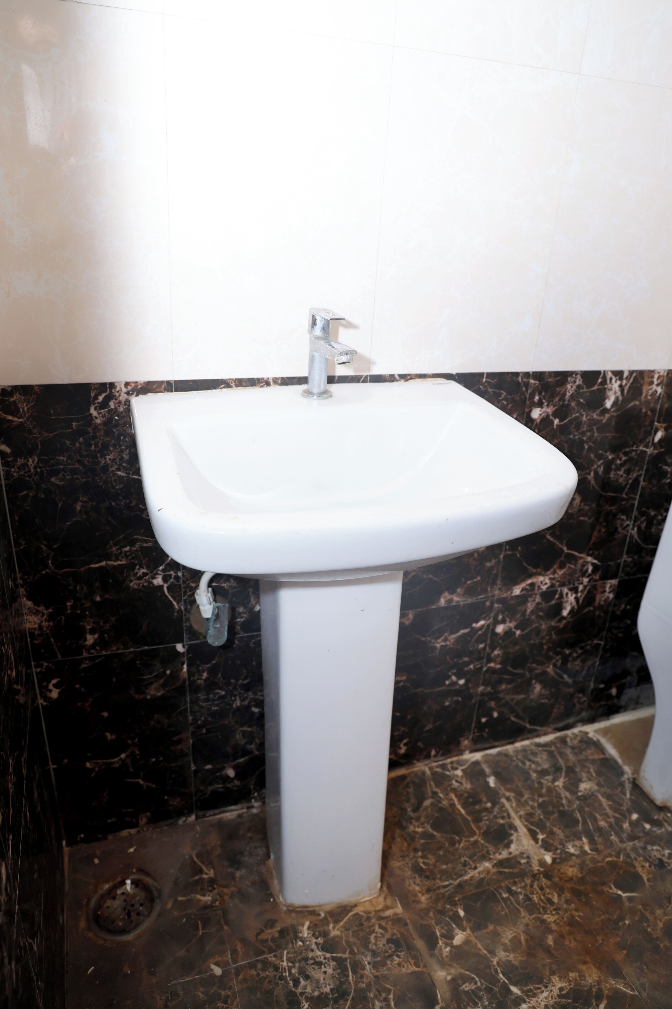 Un baño con lavamanos pedestal blanco, baldosas de mármol oscuro y desagüe visible.