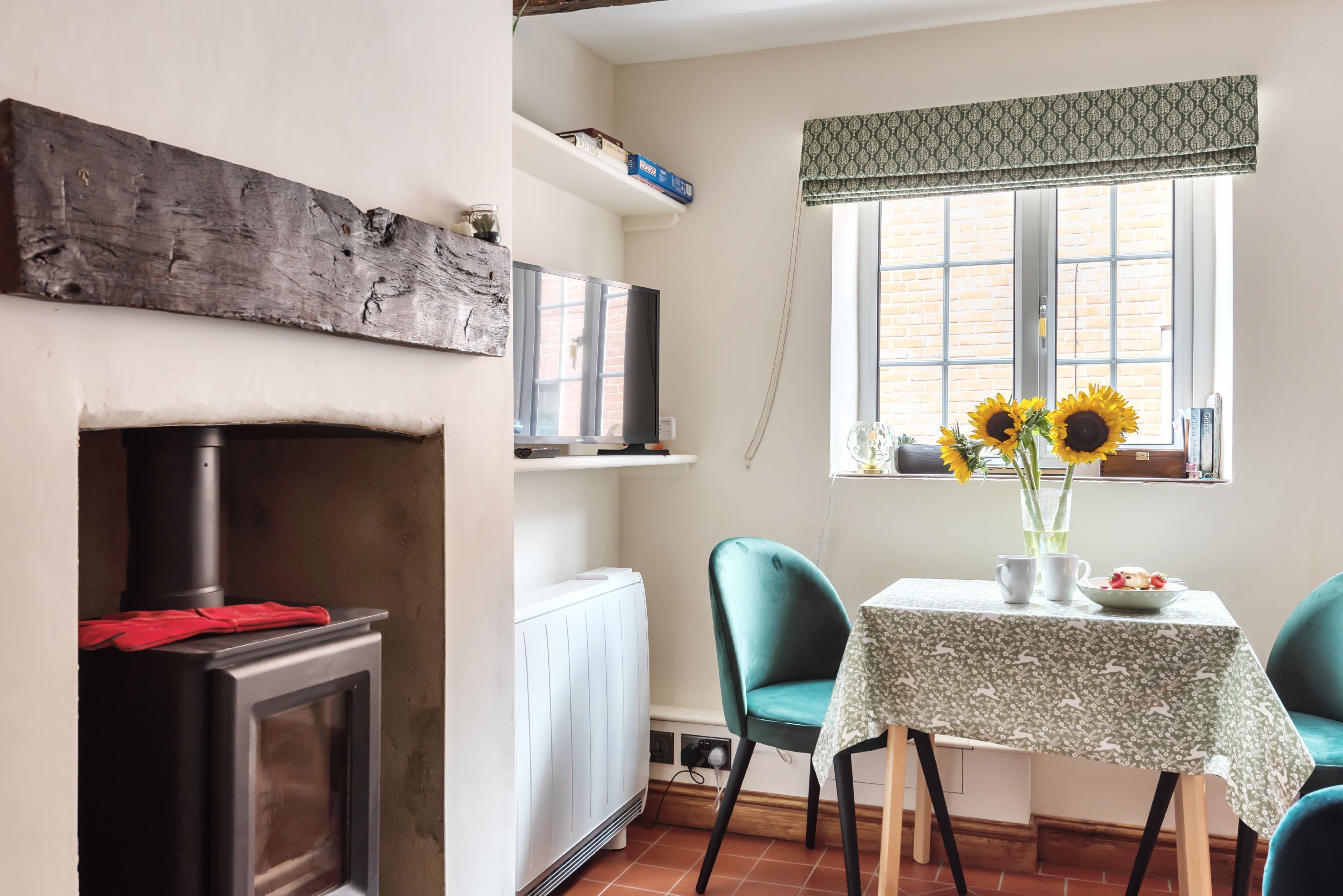 A cozy room with a fireplace, a table with a floral tablecloth, two chairs, a radiator, and a window with a patterned valance.