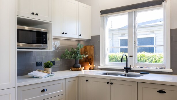 Een keuken met witte kastjes, een magnetron, een spoelbak en een raam met uitzicht op groen.