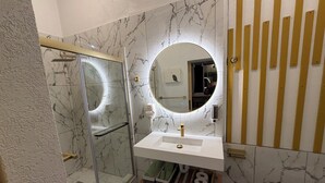 Ein Badezimmer mit Duschkabine aus Glas, einem weißen Waschbecken mit goldfarbenem Wasserhahn, einem runden Spiegel und Wänden mit Marmoroptik.