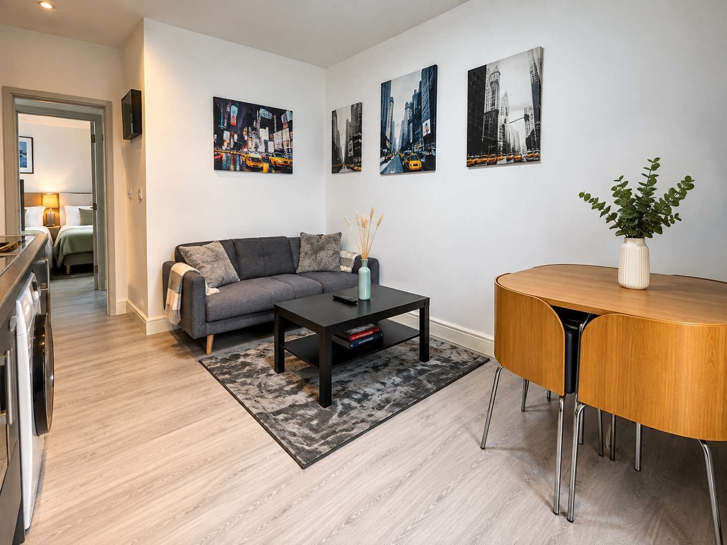 A modern living room with a sofa, coffee table, and a dining table with chairs. There are three paintings on the wall depicting cityscapes.