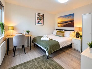 Hotellihuone, jossa on sänky, työpöytä, tuoli, yöpöytä, lamppu, taulu seinällä ja näkymä auringonlaskuun.