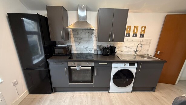 A compact kitchen with a built-in oven, microwave, and washing machine, featuring grey cabinetry and a tiled backsplash. - Sleek 1BR Flat  Perfect Getaway (Cardiff)