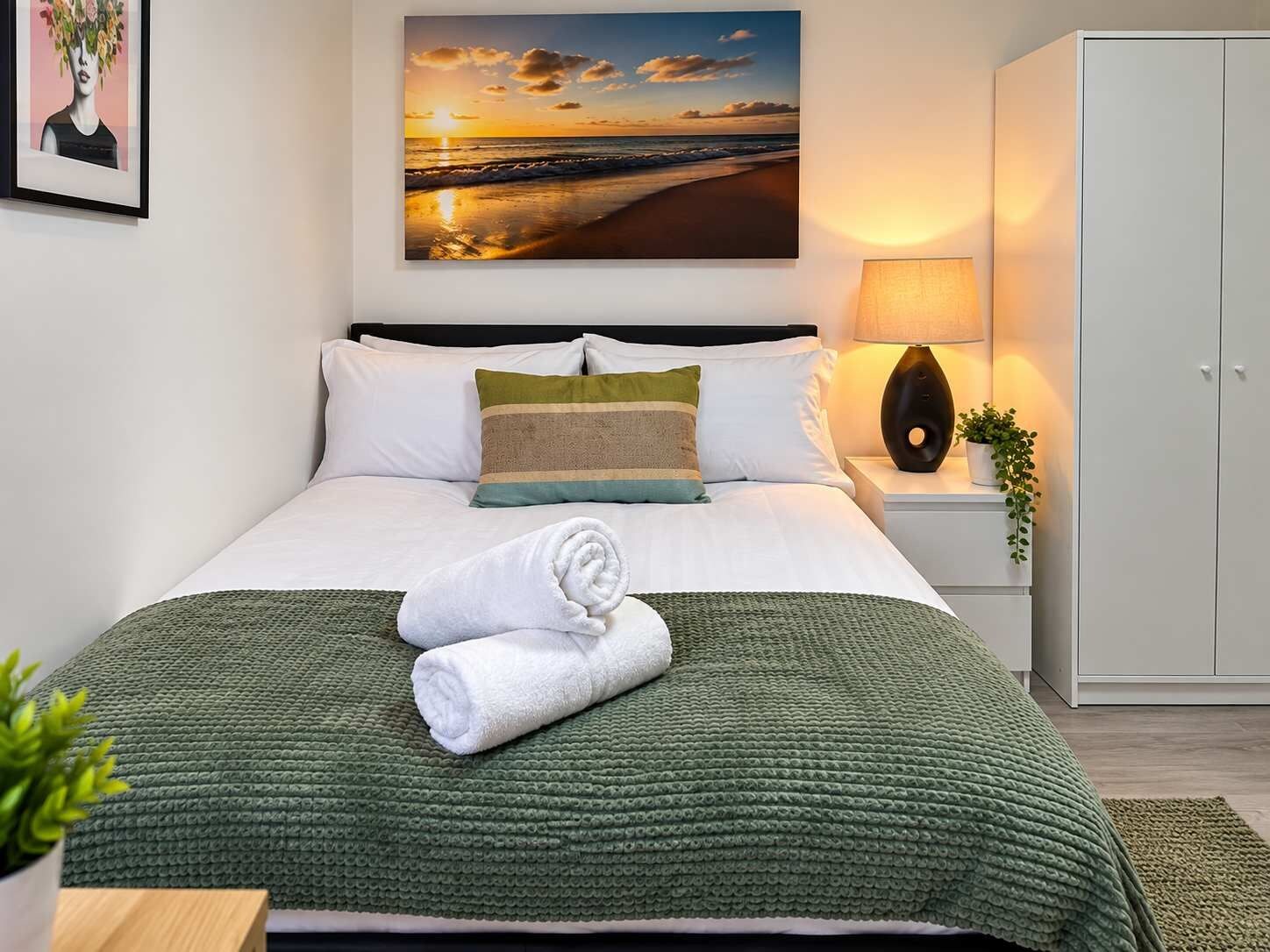 A neatly made bed with a green textured blanket, a pillow, and a rolled white towel.