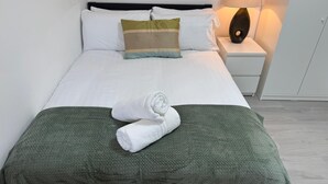 A neatly made bed with white linens, a green blanket, and a rolled towel on the bed. There is a bedside lamp on the nightstand. - Sleek 1BR Flat  Perfect Getaway (Cardiff)