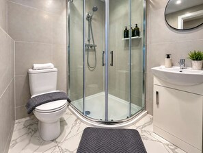 Moderni kylpyhuone, jossa on lasiseinällä varustettu suihku, wc, peilillä varustettu pesuallas ja hygieniatuotteet hyllyllä.