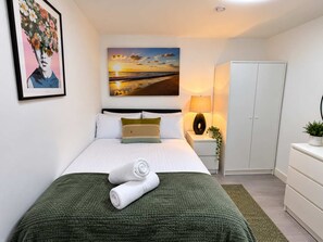 A neatly made bed with a green blanket, a painting of a sunset, a lamp, and a wardrobe. - Cardiff 1-Bed Ground-Floor Flat | Private Garden & Sofa Bed (Cardiff)