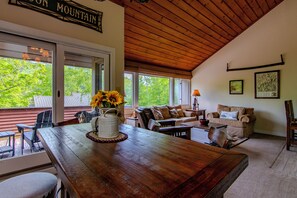 Ein Holztisch mit Sonnenblumen in einer Vase, ein Sofa und ein Sessel in einem Zimmer mit geneigter Decke und großen Fenstern.