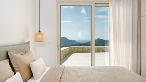 Een slaapkamer met een bed, kussens, een hanglamp en uitzicht op bergen door een glazen deur.