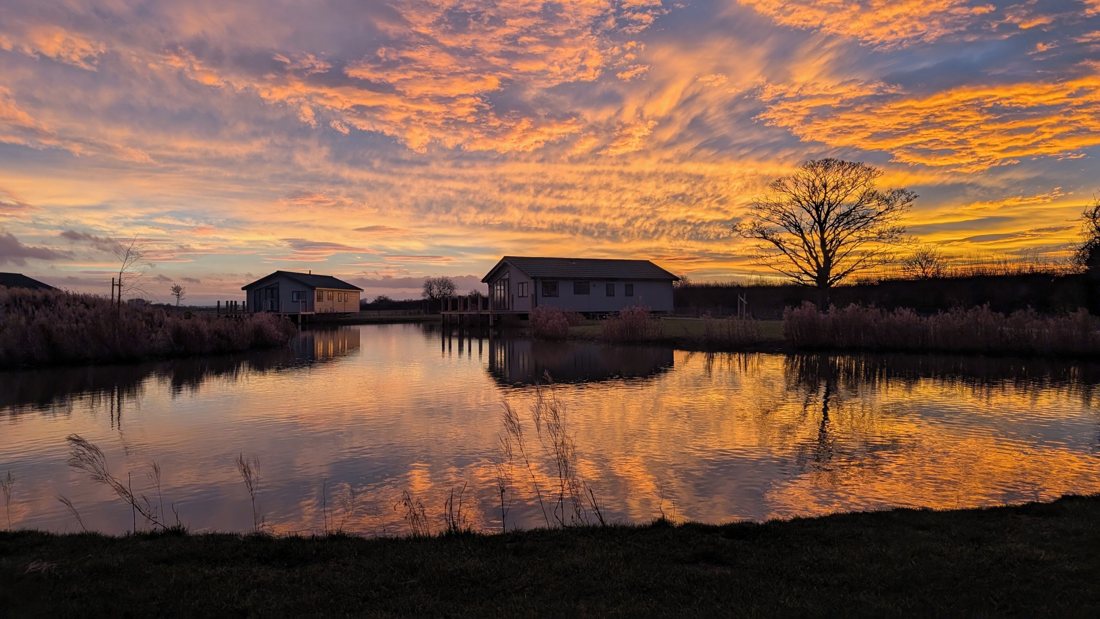 A tranquil lakeside scene with two houses, calm water, and a sunset sky.