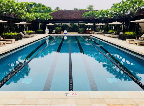 A swimming pool with clear lanes, surrounded by lounge chairs and umbrellas, and a covered area with a tiled roof.