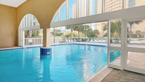 Una piscina interna con pareti in vetro trasparente e vista su palme e edifici circostanti.