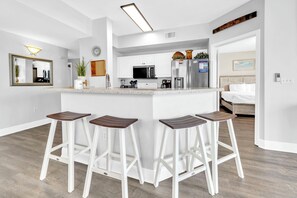 A modern kitchen with white cabinets, a central island with bar stools, a microwave, and a refrigerator. - ☀️Gulf Front Corner Views | Pools | Celadon 409-Champagne Sunsets☀️ (Panama City Beach)