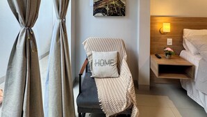A room with a bed, a chair with a 'HOME' pillow, a wall-mounted picture of the Eiffel Tower, a nightstand with a lamp, and a curtain. - Ahava Flat - Hospedagem Inteligente (Montes Claros)