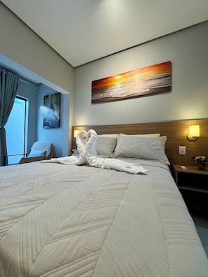Habitación de hotel con una cama grande, un cuadro de un atardecer, una silla y una mesita de noche con una lámpara.