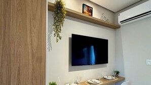 Ein moderner Essbereich mit einem langen Holztisch, weißen Stühlen, einem Wand-Fernseher und einer hängenden Pflanze.