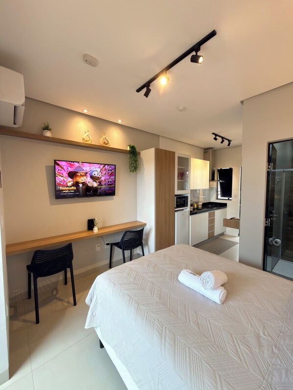 Une chambre d’hôtel moderne équipée d’un lit, d’un bureau, de chaises, d’une télévision, d’une kitchenette et d’une douche en verre.