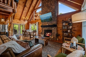 A cozy cabin interior with a stone fireplace, wooden furniture, and a large window overlooking greenery. - Resort Perks|Amazing Location|Hot Tub|Game Room (Pigeon Forge)