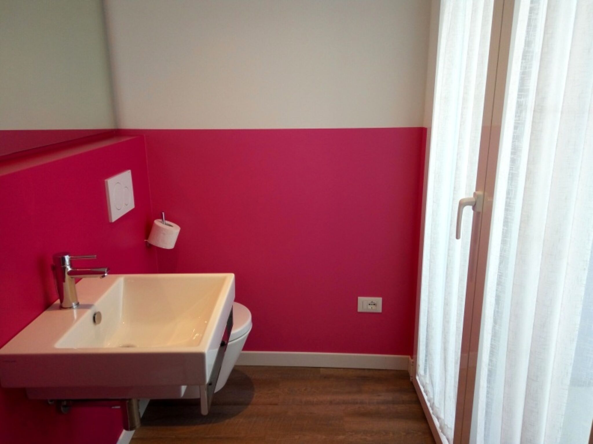 A bathroom with a white sink, pink walls, and a window with white curtains.