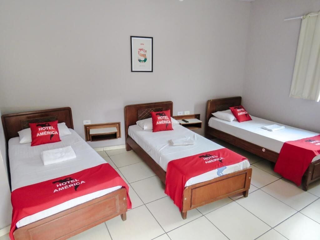 A hotel room with three beds, each with a red 'Hotel América' pillow and blanket. There is a framed picture on the wall and a window with a curtain.