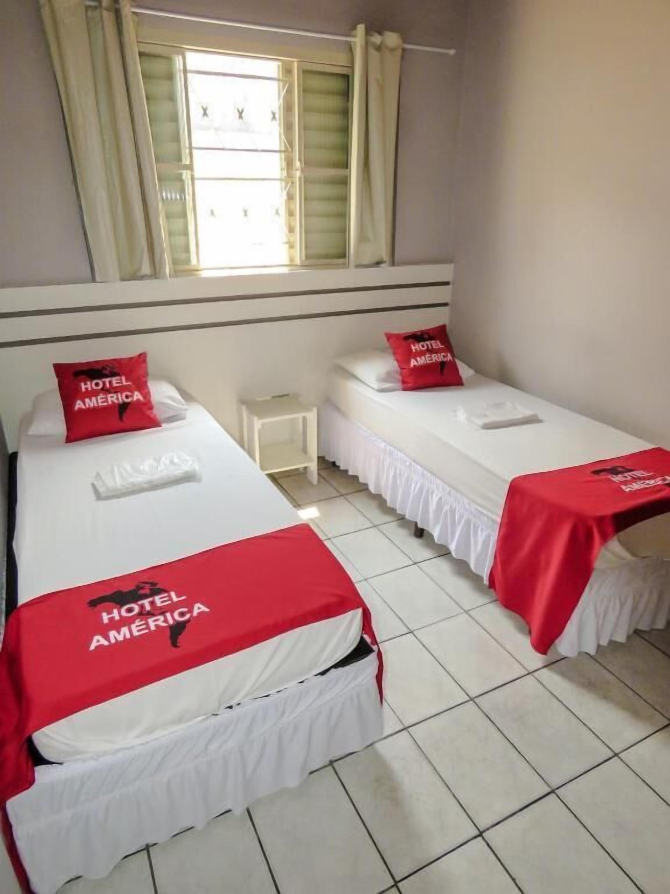 A hotel room with two beds, each with a red Hotel América pillow and bedspread. There is a small bedside table between the beds.