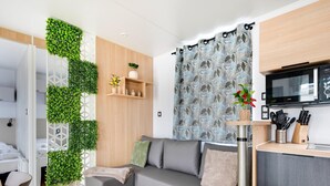 A modern living room with a grey sofa, a small table with a plant, a kitchen area with a microwave, and a decorative green wall. - Cozy Caravan in Berlare for 6 People (Berlare)