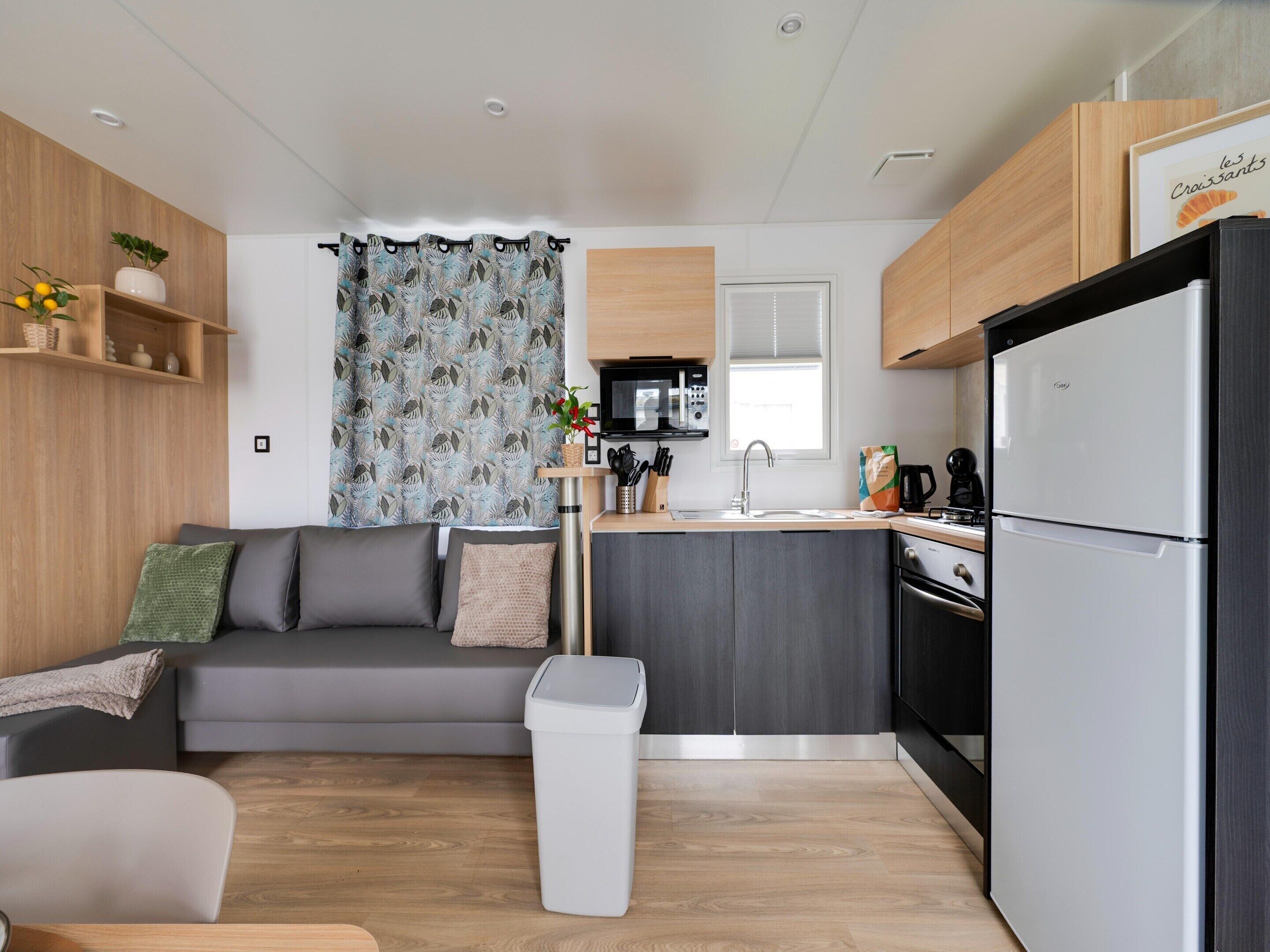 A compact living space with a kitchenette, a grey sofa, a trash bin, and a window with a curtain.