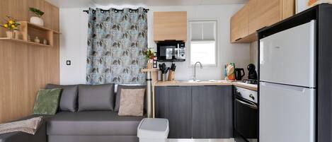 A compact living space with a kitchenette, a grey sofa, a trash bin, and a window with a curtain.
