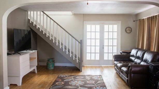 A living room with a staircase, a leather sofa, a TV on a white cabinet, and a patterned rug. - 3 Bedroom Main Level house located half an hour from Waterton. (Cardston)