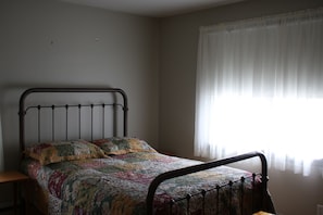 Una cama individual con estructura metálica, colcha y una ventana con cortinas traslúcidas.