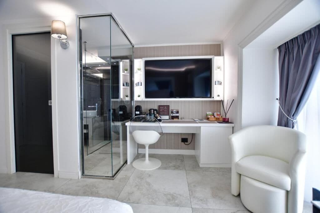 Una habitación de hotel moderna con un separador de vidrio, un televisor de pantalla plana, un escritorio con una silla y un amplio ventanal con cortinas.