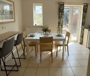 Un comedor con una mesa blanca dispuesta para cuatro, baldosas hexagonales en el suelo, un radiador y una ventana con cortinas.