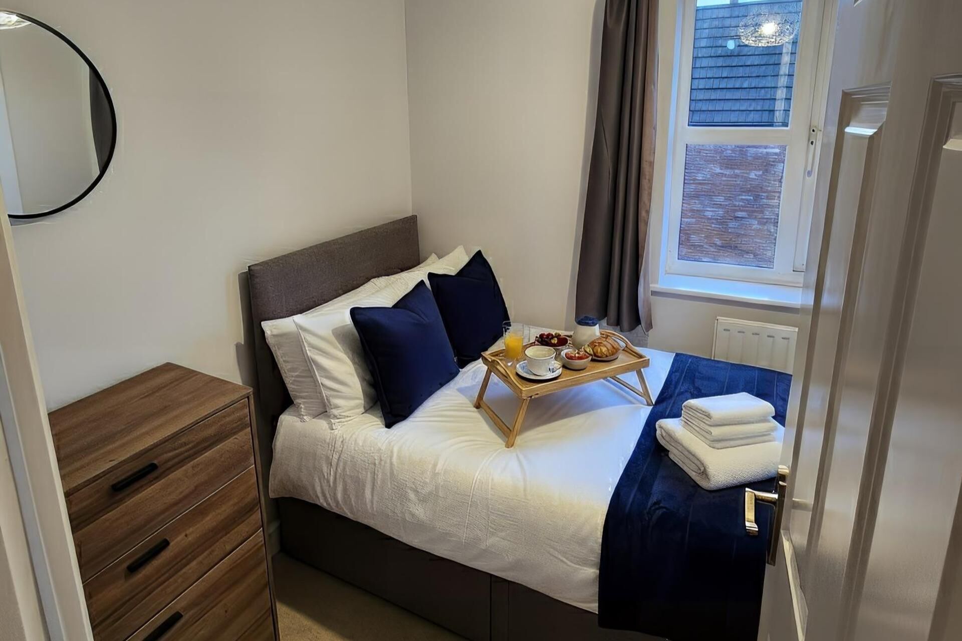 A neatly made bed with a tray of breakfast items, a bedside table with a lamp, and a window with curtains.