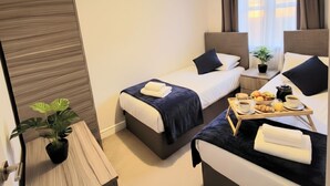 Ein Hotelzimmer mit zwei Betten, einem kleinen Tisch mit Frühstücksgebäck, einer Zimmerpflanze und einem Fenster mit Vorhängen.