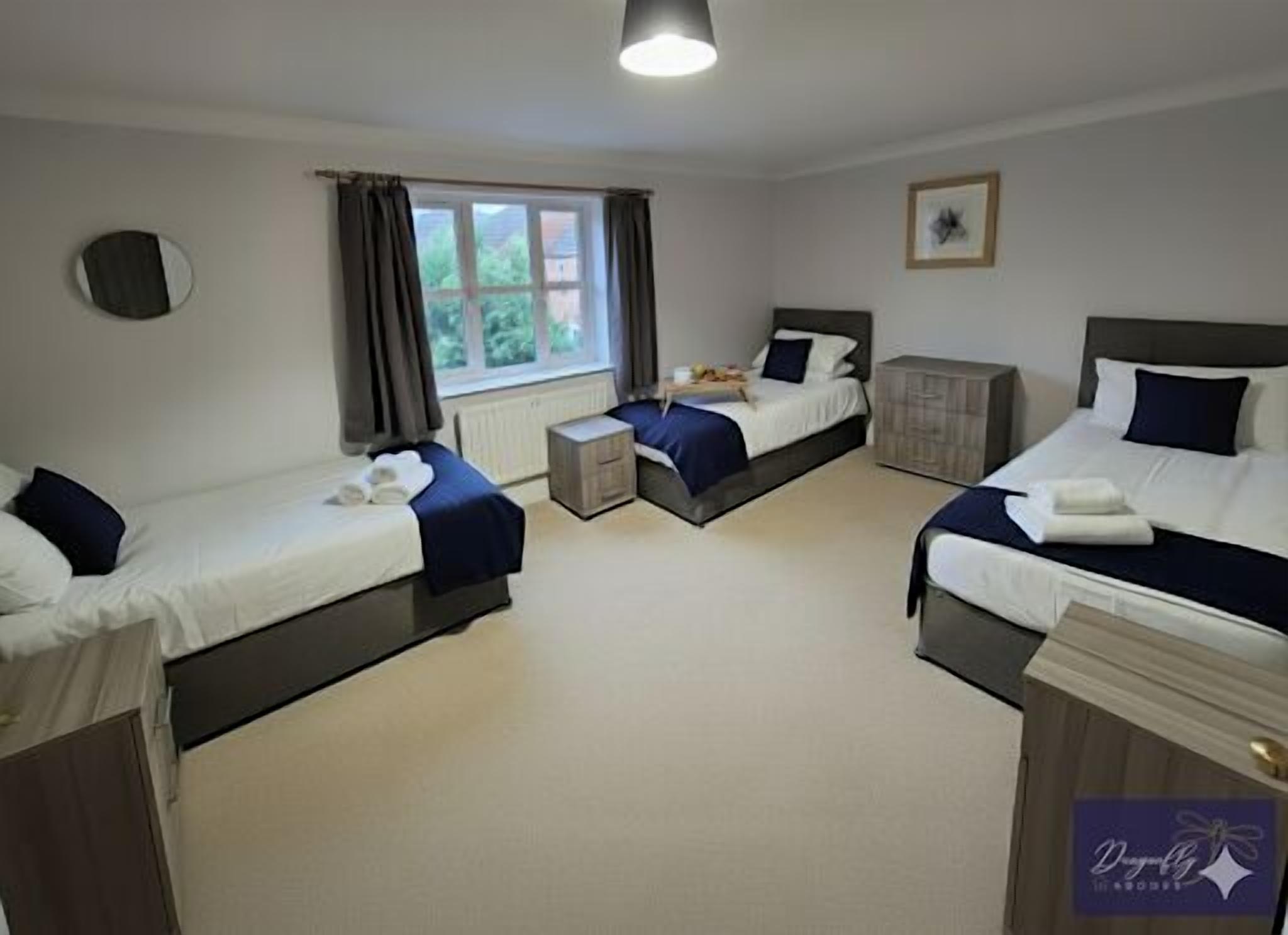 A hotel room with three single beds, a small table, a cabinet, a window with curtains, and a framed picture on the wall.