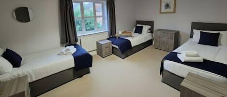 A hotel room with three single beds, a small table, a cabinet, a window with curtains, and a framed picture on the wall.