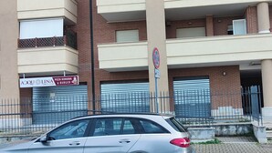 Um edifício com vários andares, uma entrada com portão metálico fechado, um letreiro de restaurante de pizza e hambúrguer, e um carro estacionado na frente.