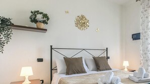 Cama bem arrumada com estrutura metálica, duas mesas de cabeceira com luminárias, uma pequena prateleira com uma planta e um objeto decorativo na parede.