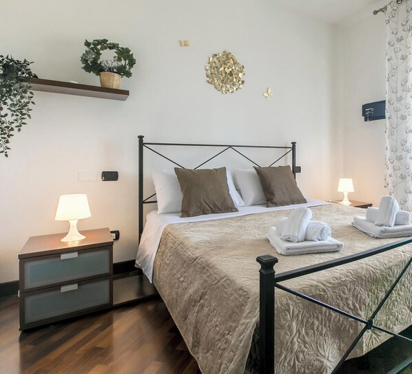 Un lit bien fait, avec un cadre en métal, deux tables de chevet avec des lampes, une petite étagère avec une plante et un élément décoratif mural.