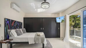 Un dormitorio moderno con una cama, televisor, ventilador de techo y vistas a palmeras.