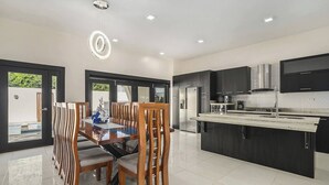Cocina moderna con armarios oscuros, una isla central y una zona de comedor con mesa de vidrio y sillas de madera.