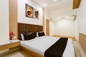A modern hotel room with a large bed, a bedside table, a nightstand, a wall-mounted light, and a decorative vase with flowers.