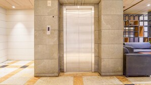 An elevator in a lobby with tiled flooring and a seating area with a sofa and ottoman. - Deluxe Ramora Grand (Thindukkal)