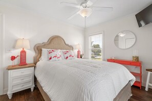 A bedroom with a bed, nightstands, a dresser, a ceiling fan, a window with blinds, and a wall-mounted TV.