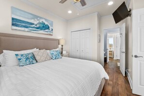 A bedroom with a large bed, a ceiling fan, a mounted TV, and a view into the adjacent bathroom.