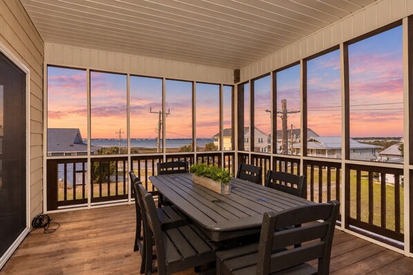 A covered outdoor area with a table and chairs, large windows offering a view of the sea and houses, and a wooden deck.