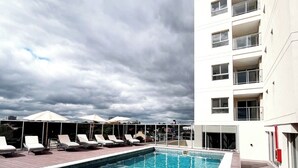 A swimming pool with lounge chairs, a covered seating area, and a multi-story building with balconies.